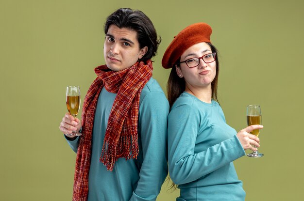 Disgustado mirando cámara pareja joven en el día de San Valentín chico con bufanda chica con sombrero sosteniendo una copa de champán aislado sobre fondo verde oliva