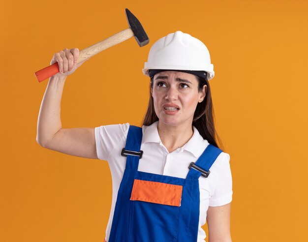 Disgustado mirando a la mujer joven constructor de lado en uniforme sosteniendo un martillo aislado en la pared naranja