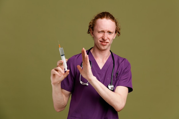 disgustado mostrando gesto joven médico masculino vistiendo uniforme con estetoscopio sosteniendo jeringa aislado sobre fondo verde