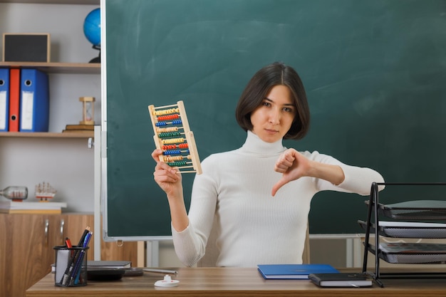 disgustado mostrando los pulgares hacia abajo joven maestra sosteniendo abacus sentado en el escritorio con herramientas escolares en el aula