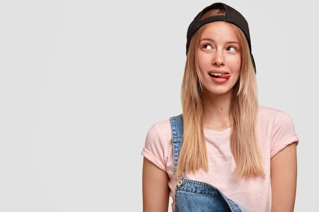 Disparo aislado de una adolescente bonita de moda mira a un lado con expresión soñadora, muestra la lengua, usa gorra negra y un mono de moda, contempla algo agradable, se encuentra en el interior