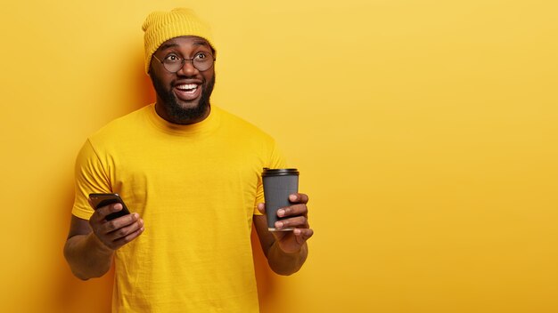 Disparo aislado de feliz adolescente afroamericano de buen humor, verifica el buzón de correo electrónico en Internet, utiliza un dispositivo moderno, bebe café para llevar, sonríe positivamente, posa sobre una pared amarilla. Tiempo libre