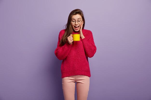 Disparo aislado de feliz mujer llena de alegría mira una taza de bebida aromática caliente, sostiene una taza amarilla, usa gafas, un puente rojo, se encuentra contra la pared púrpura. Mujer alegre tiene descanso para tomar café. Concepto de bebida