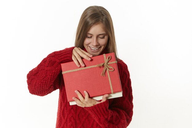 Disparo aislado de una hermosa joven con elegantes gafas y un jersey marrón sosteniendo una caja abierta con un regalo de cumpleaños, con una expresión facial triste y decepcionada, no le gusta lo que hay dentro