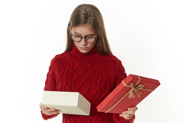 Disparo aislado de una hermosa joven con elegantes gafas y un jersey marrón sosteniendo una caja abierta con un regalo de cumpleaños, con una expresión facial triste y decepcionada, no le gusta lo que hay dentro