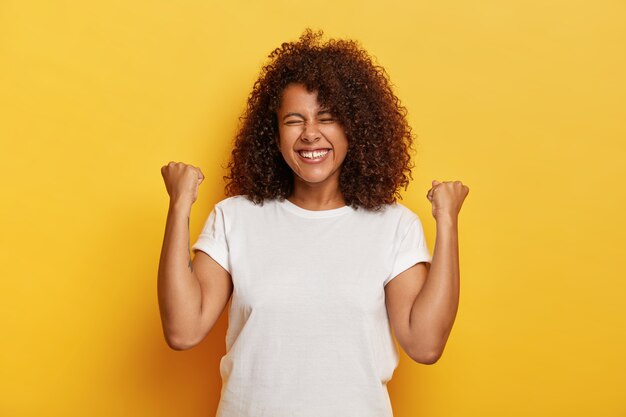 Disparo aislado de hermosa mujer exitosa con cabello rizado, levanta los puños cerrados, celebra el triunfo, está muy complacido y feliz, cierra los ojos de placer, usa camiseta blanca. ¡Sí, lo hice!