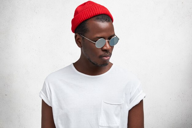 Disparo aislado de hombre afroamericano joven serio de moda en gafas de sol, sombrero rojo de moda y camiseta casual, mira a un lado, aislado sobre estudio blanco