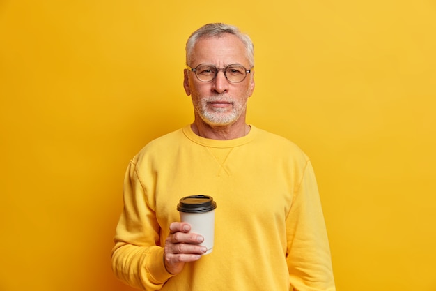 Disparo aislado de hombre guapo con barba sostiene café para llevar desechable y mira seriamente al frente tiene descanso vestido con poses de puente brillante contra la pared amarilla