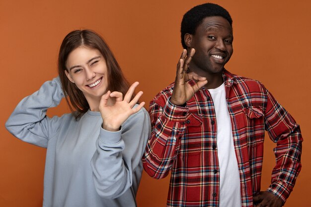 Disparo aislado de una joven europea encantadora y positiva con una linda sonrisa amplia posando con su atractivo y alegre novio negro, ambos guiñando un ojo y mostrando buenos gestos