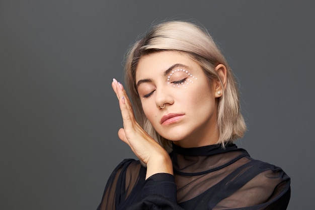 Disparo aislado de una linda joven encantadora de veinte años en una blusa negra transparente manteniendo los ojos cerrados y tocando suavemente su piel suave con un anillo en la nariz, un elegante corte de pelo y un maquillaje brillante