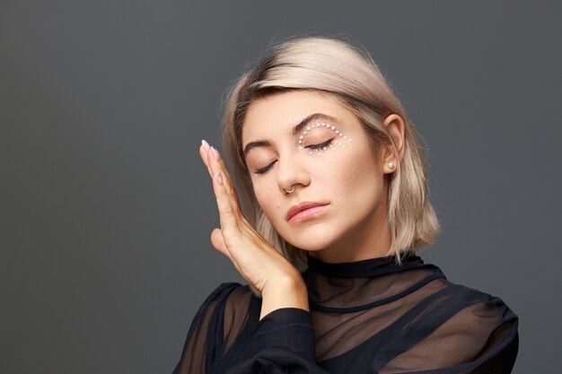 Disparo aislado de una linda joven encantadora de veinte años en una blusa negra transparente manteniendo los ojos cerrados y tocando suavemente su piel suave con un anillo en la nariz, un elegante corte de pelo y un maquillaje brillante