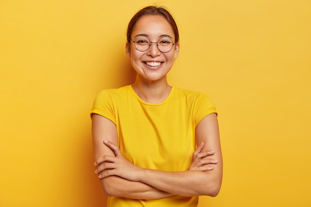 Disparo aislado de mujer alegre complacida con apariencia oriental, sonríe ampliamente, está de buen humor, entretenida por amigos divertidos, vestida de manera informal, usa grandes lentes transparentes, aislados en amarillo