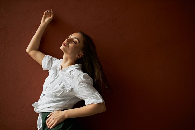 Disparo aislado de una mujer atractiva sensual con el pelo largo y castaño suelto cerrando los ojos en el disfrute, recostándose en la pared del estudio del espacio de copia en blanco, con mirada complacida, levantando en la mano, vistiendo camisa con