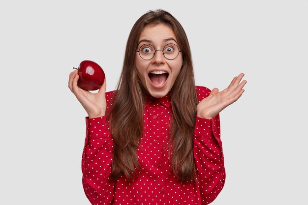 Disparo aislado de mujer atractiva tiene expresión alegre, sostiene la manzana roja en una mano, expresa sentimientos positivos, viste camisa formal