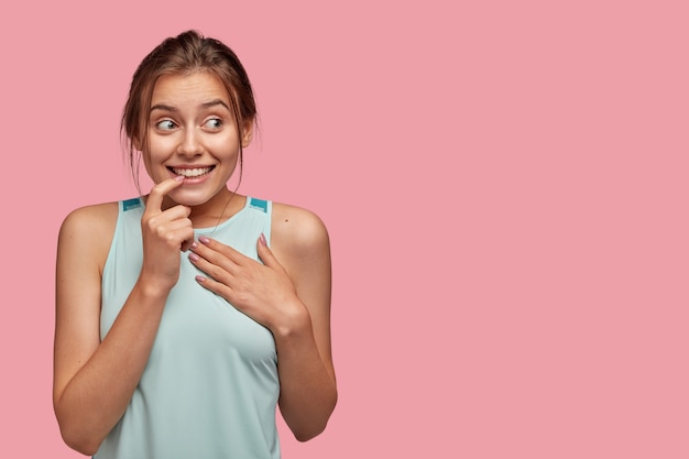 Disparo aislado de mujer europea positiva muerde el dedo, mantiene la mano en el pecho, sonríe positivamente, mira a un lado, viste ropa casual, escucha información intrigante, aislado sobre una pared rosa con espacio libre