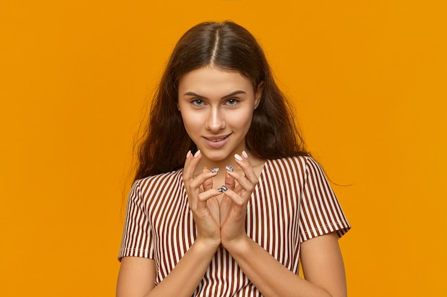 Foto gratuita disparo aislado de mujer joven astuta inteligente con un vestido elegante con rayas verticales entrelazando las manos