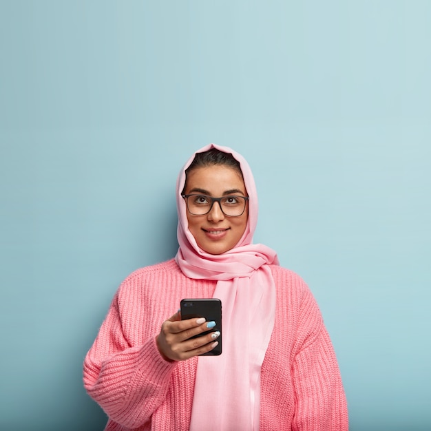 Disparo aislado de mujer musulmana bastante soñadora utiliza el teléfono celular para enviar mensajes en las redes sociales, enfocado hacia arriba, piensa en el contenido del mensaje, se para sobre la pared azul.