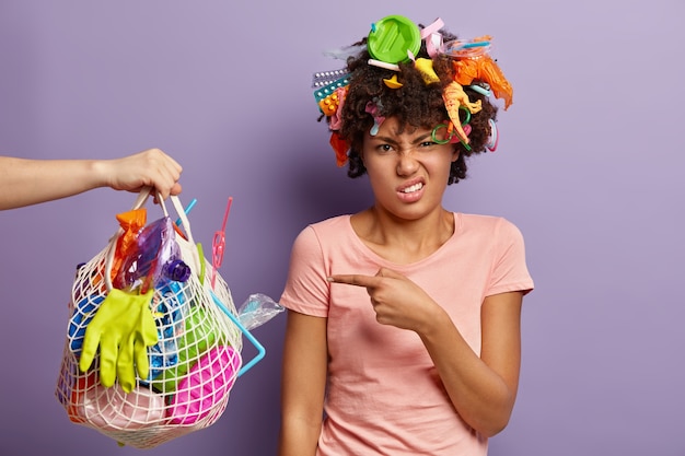 Foto gratuita disparo aislado de una mujer de piel oscura desesperada apunta a una bolsa de basura que lleva a una persona desconocida, ocupada en el día mundial del medio ambiente, vestida con una camiseta informal, se encuentra sobre una pared púrpura, recoge basura