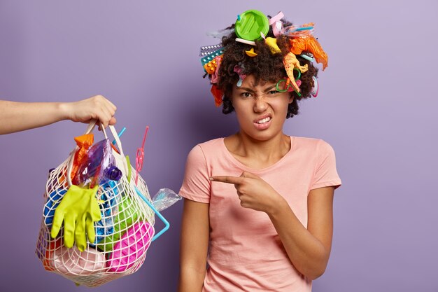 Disparo aislado de una mujer de piel oscura desesperada apunta a una bolsa de basura que lleva a una persona desconocida, ocupada en el Día Mundial del Medio Ambiente, vestida con una camiseta informal, se encuentra sobre una pared púrpura, recoge basura