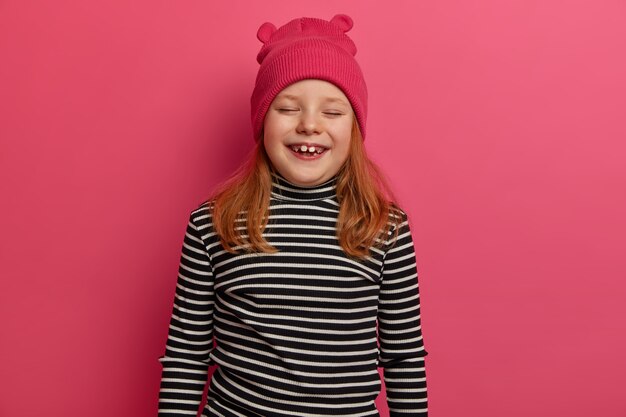 Disparo aislado de una niña pelirroja sonríe y se ríe positivamente, viste un sombrero rosa y un jersey a rayas, siendo muy emocional, viene en la fiesta de cumpleaños, aislada en la pared rosa. Concepto de emociones felices