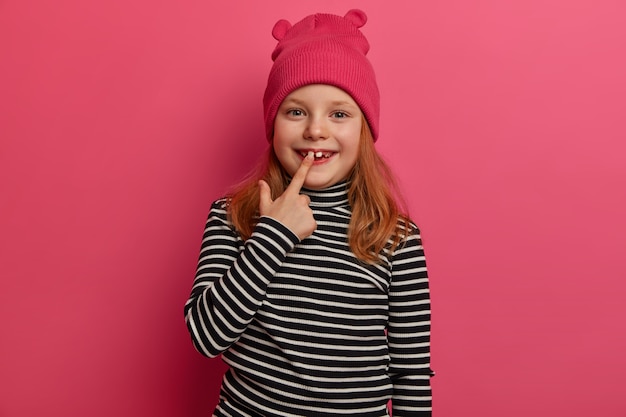 Disparo aislado de un niño pequeño que se regocija con un nuevo diente adulto, tiene el pelo rojo, vestido con un suéter a rayas y un elegante sombrero, tiene una encantadora expresión positiva posa sobre una pared rosada. Mira mis dientes blancos