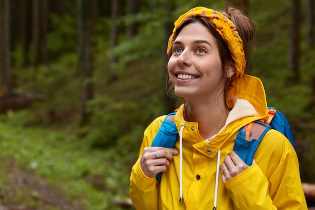 Foto gratuita disparo al aire libre de una joven alegre que mira pensativamente a la distancia, lleva una diadema amarilla y un impermeable, deambula por el bosque