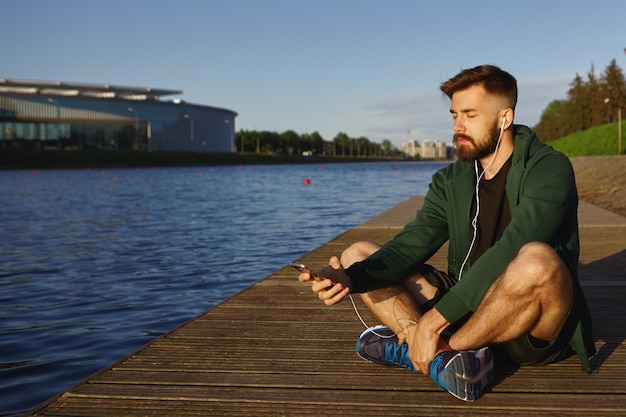 Disparo al aire libre de un joven guapo sin afeitar con rastrojo pasando una tranquila mañana de verano solo junto al lago, sentado con los ojos cerrados, escuchando pistas de música meditativa en un teléfono inteligente moderno