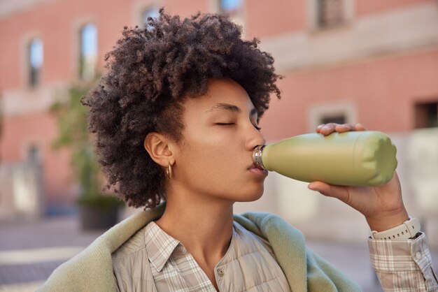 Disparo al aire libre de la joven de pelo rizado mantiene los ojos cerrados mientras bebe agua fresca de la botella se toma un descanso después del entrenamiento