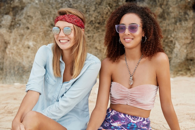 Disparo al aire libre de modelos femeninas relajadas y despreocupadas de diferentes nacionalidades sentadas en la playa de arena contra el acantilado, estando de buen humor mientras disfrutan de la unión y el verdadero amor mutuo entre sí