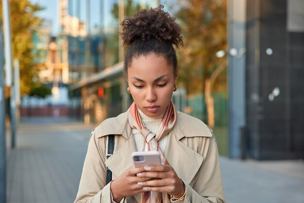 Disparo al aire libre de una mujer joven de aspecto agradable hace banca a través de la aplicación tiene un teléfono móvil moderno navega por las redes sociales