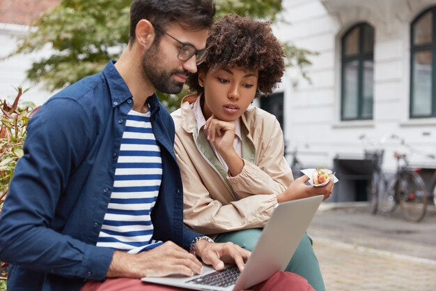 Disparo al aire libre de pareja de raza mixta tiene miradas atentas en la computadora portátil