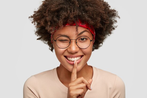 Disparo en la cabeza de la alegre joven mujer afro juguetona hace gesto de silencio con expresión positiva, parpadea el ojo