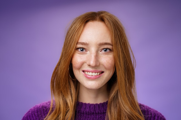 Disparo en la cabeza de la encantadora mujer joven pelirroja atractiva y feliz con pecas y sonrisa blanca brillante, sonriendo satisfecho a la cámara como posando sobre fondo púrpura amistoso y encantado.