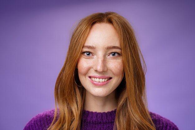 Disparo en la cabeza de la encantadora mujer joven pelirroja atractiva y feliz con pecas y sonrisa blanca brillante, sonriendo satisfecho a la cámara como posando sobre fondo púrpura amistoso y encantado.