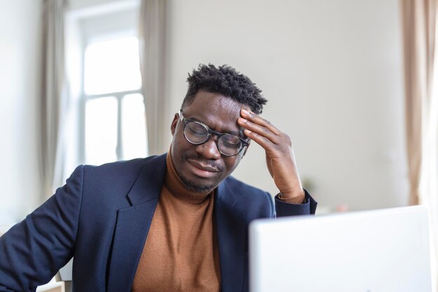 Disparo en la cabeza estresado joven afroamericano tocando la frente que sufre de un terrible dolor de cabeza trabajando en la computadora en la oficina de casa Frustrado confundido hombre birracial que tiene sentimientos dolorosos en la cabeza