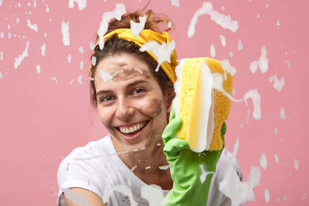 Disparo en la cabeza de feliz hermosa ama de casa joven positiva con encantadora sonrisa, lavar la ventana en la cocina, limpiar la espuma gruesa de la superficie del vidrio, disfrutar del proceso de limpieza, sonriendo