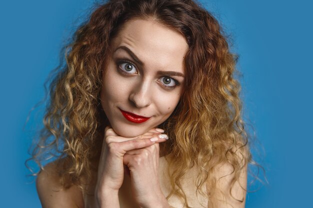 Disparo en la cabeza de una hermosa chica elegante con cabello voluminoso y labios rojos con mirada sospechosa, levantando las cejas y tomados de la mano debajo de la barbilla. Linda mujer mirando con expresión juguetona