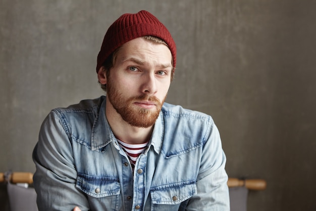 Disparo en la cabeza del hipster masculino con barba elegante con sombrero y camisa de jeans que mira con dudosa expresión indecisa, se siente frustrado y desconcertado por tomar una decisión importante