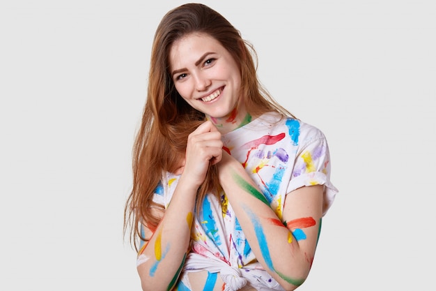 Disparo en la cabeza de una joven modelo positiva que mantiene las manos juntas, sonríe suavemente, usa una camiseta manchada informal, le gusta pintar