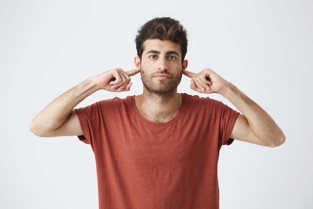 Disparo en la cabeza del molesto chico italiano elegante con camiseta roja que parece gruñón, cerrando las orejas con los dedos mostrando falta de respeto a sus palabras de amigo.