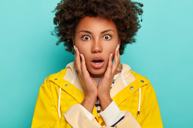 Disparo en la cabeza de una mujer afro asombrada que no puede creer en noticias impactantes, se toca las mejillas con las palmas, abre la boca de gran sorpresa, vestida con impermeable amarillo, expresa miedo y desconcierto