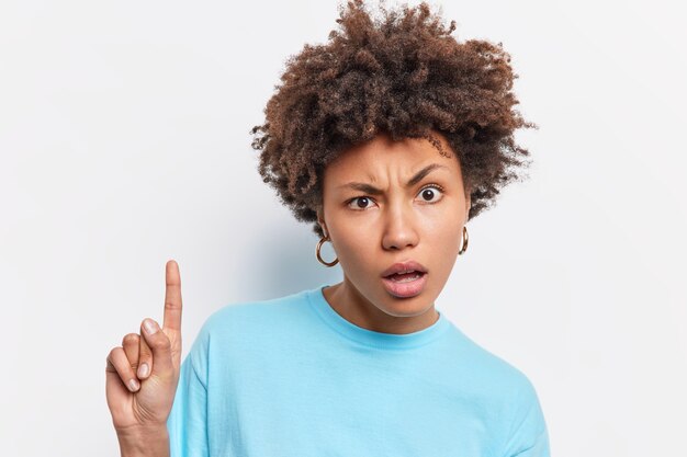 Disparo en la cabeza de una mujer afroamericana de pelo rizado disgustada que parece desconcertada, indica que el dedo índice de arriba muestra algo increíble vestido con una camiseta azul casula aislada sobre una pared blanca