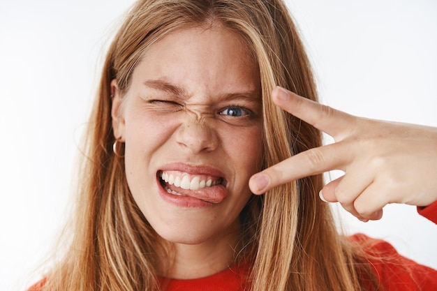 Disparo en la cabeza de una mujer despreocupada joven divertida, emotiva y carismática que hace muecas sacando la lengua mostrando un gesto de paz y un guiño expresando emociones positivas y emocionadas posando sobre una pared gris