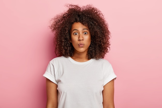 Disparo en la cabeza de una mujer encantadora con piel oscura, mantiene los labios doblados, tiene un corte de pelo afro, hace muecas, tiene belleza natural, sin maquillaje, viste una camiseta blanca informal, aislada en una pared rosa