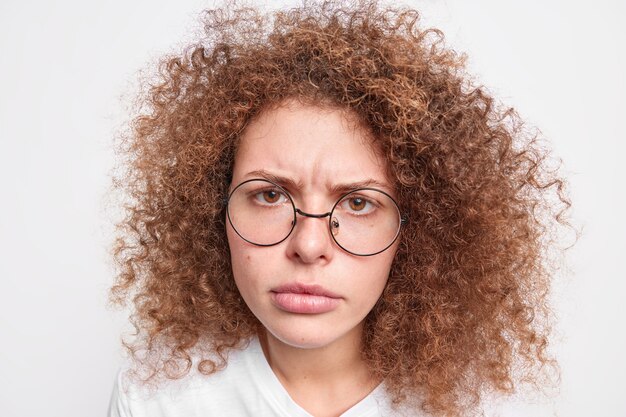 Disparo en la cabeza de una mujer europea joven molesta enojada con el pelo rizado y tupido que se ve infelizmente lleva gafas redondas aisladas sobre una pared blanca. Concepto de reacciones y expresiones humanas.