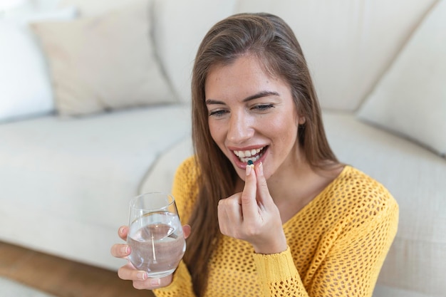 Disparo en la cabeza mujer joven sonriente sosteniendo una píldora y un vaso de agua pura fresca Señora milenaria saludable tomando medicina antioxidante vitaminas suplementos de belleza para el cabello piel uñas concepto de salud