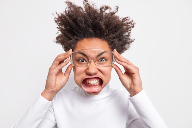 Disparo en la cabeza de una mujer de pelo rizado irritada que aprieta los dientes, una sonrisa de suficiencia, exclama de ira, usa lentes transparentes, cuello alto blanco, posa en el interior, siendo súper loco y muy enojado con alguien.