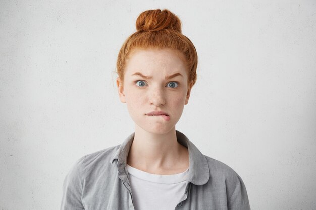 Disparo en la cabeza de una niña caucásica pecosa pelirroja ansiosa levantando las cejas y mordiéndose los labios inferiores con mirada asustada, nerviosa o enojada, esperando algo con impaciencia, posando aislado