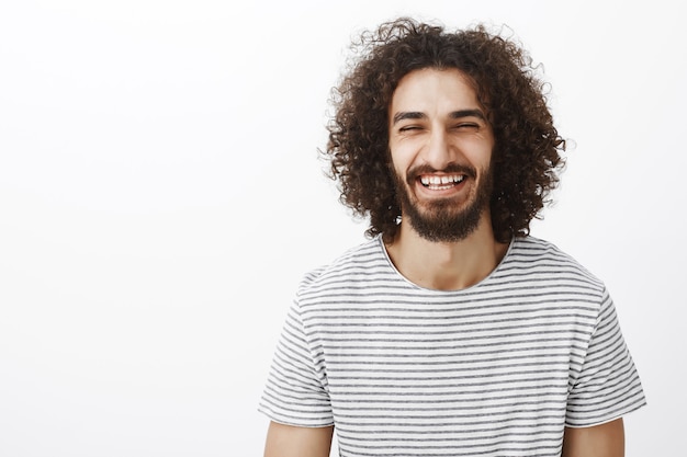 Foto gratuita disparo de cintura para arriba del amable chico hispano barbudo feliz con el pelo rizado, vistiendo elegante camiseta a rayas, riendo a carcajadas con alegría