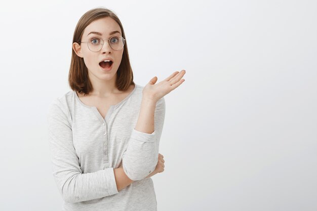 Disparo de cintura para arriba de una chica inteligente, guapa, habladora, entretenida y divertida con gafas y blusa agitando la palma durante la conversación, de pie con la boca abierta en medio de una discusión interesante